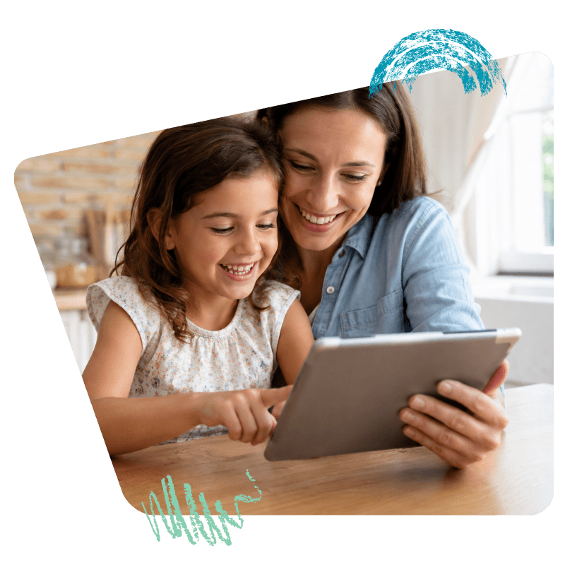 Una niña y su mamá riendo mientras usan una tablet en la mesa de la cocina